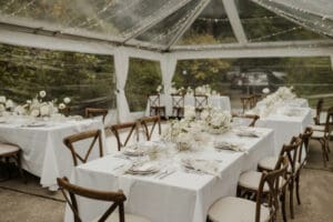 a table set with a white tablecloth and napkins with brown wooden chairs in a glass-walled and -ceilinged space
